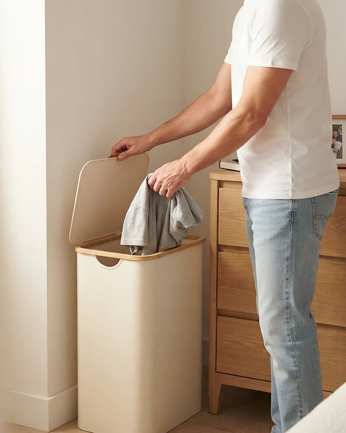 Homme qui dépose un t-shirt dans le panier à linge étroit 65 L beige, couvercle relevé, à côté d'une commode